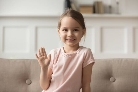 Head shot portrait little girl sitting on sofa at home wave hand look at camera use webcam online communication with friend, relative, grandparents, kid greeting make video call recording vlog conceptの写真素材