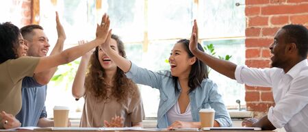 Wide horizontal image multicultural cheerful businesspeople sitting together at meeting giving high...