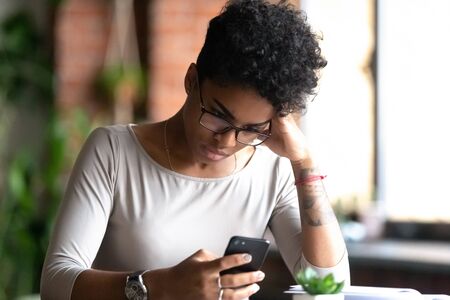 Mad african american young woman in glasses sit at desk hold cellphone annoyed with slow internet connection, angry black biracial millennial girl using smartphone having cell problems or virus attackの写真素材