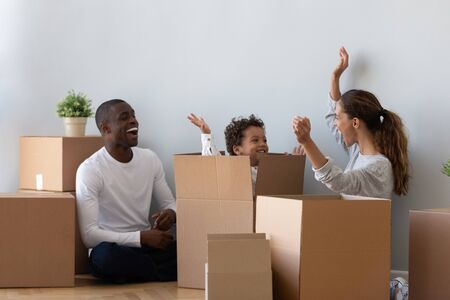 Happy mixed race ethnicity family and cute kid son playing on moving day, young parents tenants renters home owners having fun with child jump out of box unpack laughing enjoy relocation in new flatの写真素材