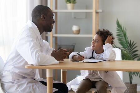 Cute african kid boy wear medical uniform play doctor talk to pediatrician at consultation, happy black paediatrician laugh meeting funny child patient pretend gp in hospital, children health careの写真素材