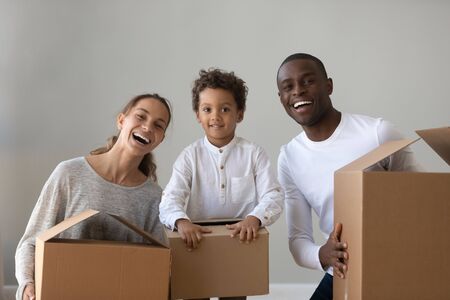 Happy african american family renters hold boxes looking at camera, smiling mixed race ethnicity parents and little child son unpack in new home apartment on moving relocation day concept, portraitの写真素材