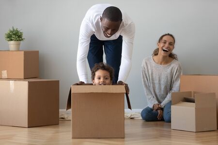 Happy black dad playing with cute little mixed race son riding in box looking at camera on moving...