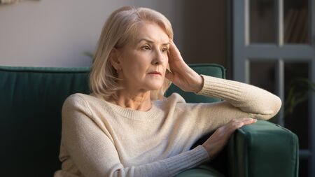 Pensive thoughtful middle aged lady looking away sit alone at home feel anxious lonely, sad depressed melancholic old mature woman suffer from sorrow grief thinking of problem suffer from solitudeの写真素材