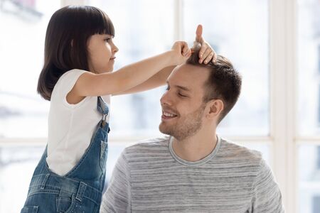 Cute funny little kid daughter playing with young single father brushing hair, small child girl holding comb pretend hairdresser doing hairstyle having fun with happy dad bonding together at homeの写真素材