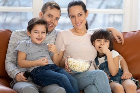 Happy family mom dad and cute little kids son daughter eat popcorn hold remote control watch tv show sit on sofa together, parents having fun lounge with children view television at home on couchの写真素材