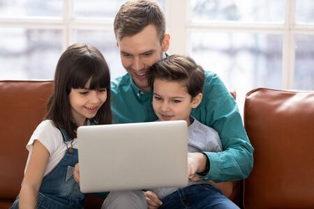 Cute small kids son daughter learning using laptop watching cartoons with happy dad, young father and children doing online shopping browsing internet on computer sit together on couch at homeの写真素材