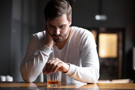 Upset young man drinker alcoholic sitting at bar counter with glass drinking whiskey alone, sad depressed addicted drunk guy having problem suffer from alcohol addiction abuse, alcoholism conceptの写真素材
