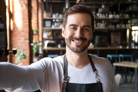 Smiling young male waiter wear apron take selfie looking at camera, head shot portrait of happy businessman entrepreneur small cafe business owner posing making photo recording video, phone cam viewの写真素材