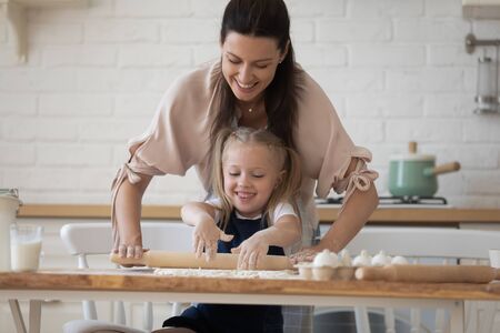 In cozy kitchen beautiful woman helping to small diligent daughter flattening dough with rolling pin...