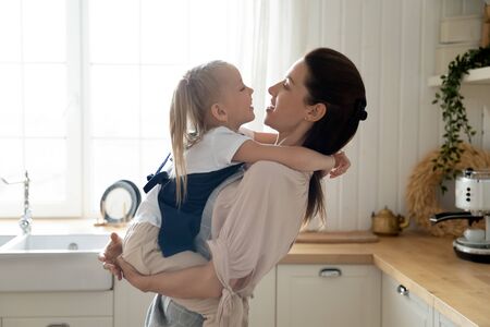 Beautiful mother holding on hands adorable preschool daughter family wearing aprons looking at each other, smiling standing side view spending time in modern cozy light kitchen after cook food togetherの写真素材