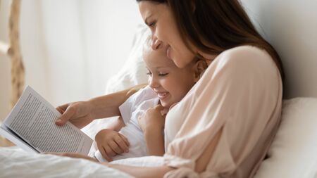 Close up loving caress mother lying with daughter in bed cuddles sweet child reading interesting storybook, preschool smiling kid girl enjoy fairytale before go to sleep good pastime and hobby conceptの写真素材