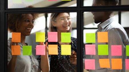 Happy friendly multicultural creative young business team people work together having fun planning project on sticky notes, three coworkers students group share ideas laugh at scrum teamwork meetingの写真素材