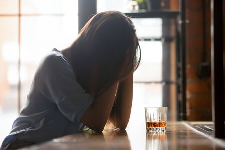 Lonely unhappy woman sitting alone with glass of whiskey, hiding face in hands, thinking about...