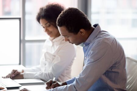 Focused african American boss at workplace write evaluate female trainee internship at company, black millennial female intern or employee look at male supervisor assess her work results or reportの写真素材