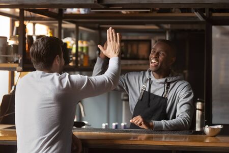Smiling African American barista giving high five with client, excited bartender wearing apron standing behind bar counter, greeting friend or regular customer in coffeehouse, celebrating successの写真素材