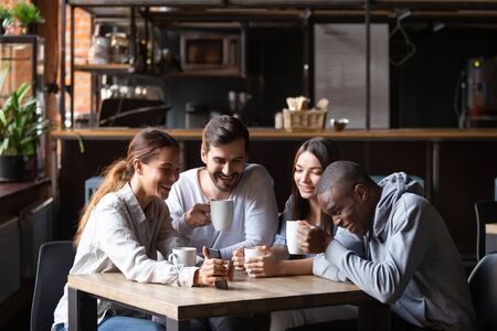 Diverse friends having fun in cafe together, looking at smartphone screen, watching funny video or...