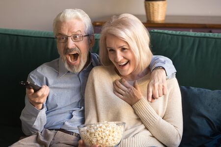 Excited elderly husband and wife watching football match on tv with popcorn, or funny comedy movie. Senior middle aged happy family couple sport fans celebrating favorite team victory.の写真素材