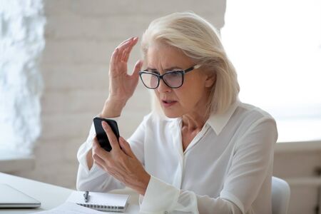 Confused senior businesswoman sit at office desk hold cellphone experience internet connection...