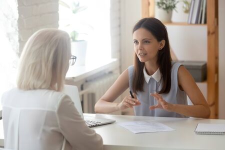 Focused millennial female talk with senior woman client consulting over contract or paper document, young recruiter or HR specialist speak with applicant or customer, have consultation in officeの写真素材