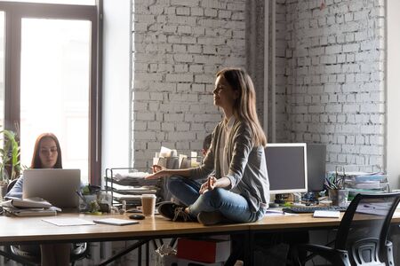 Peaceful businesswoman meditating with closed eyes on office desk, young intern, employee practicing yoga on table at work, taking pause, sitting in lotus pose at workplace, stress relief conceptの写真素材