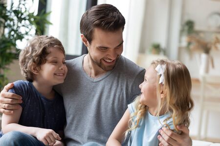 Happy young father embracing two little laughing kids siblings, joking, having fun together in living room. Adorable small children communicating with handsome daddy, enjoying weekend time at home.の写真素材