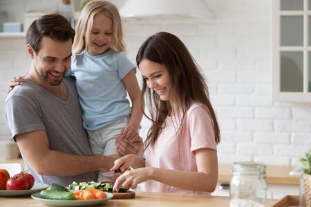 Head shot young handsome father holding little adorable happy daughter, watching smiling millennial mommy chopping fresh vegetables on wooden board, preparing healthy lunch dinner in kitchen at home.の写真素材