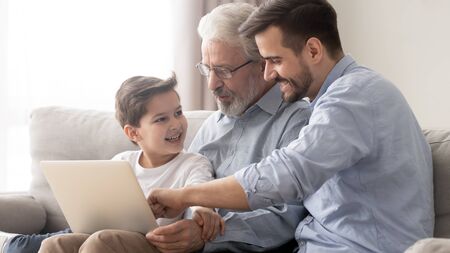 Young adult father and old senior grandfather teach little child son grandson using laptop sit on...
