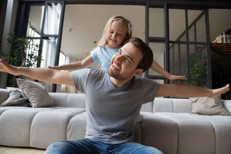 Head shot joyful young single father playing with cute small preschool daughter at modern living room. Happy little girl pretending to be plane, flying with smiling daddy, enjoying weekend time.の写真素材
