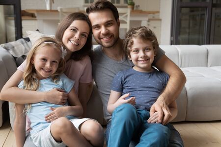 Portrait of happy full bonding family of four, sitting at warm wooden heated floor in living room at home. Smiling lovely young parents hugging little cute children siblings, looking at camera.の写真素材