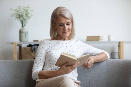 Relaxed beautiful mature woman reading book, sitting on comfortable couch, calm satisfied older female enjoying interesting literature, favorite novel or poetry, resting on weekend at homeの写真素材