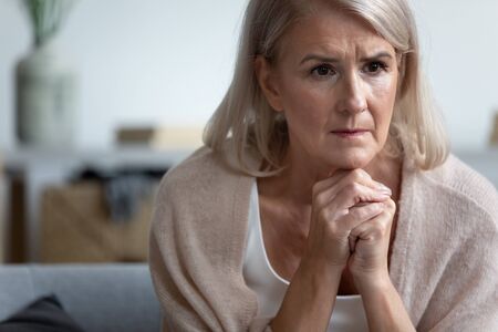 Upset thoughtful mature woman sitting alone, thinking about emotional problem, feeling unwell, lost in thoughts, dementia or mental disorder, frustrated older female looking in distance close upの写真素材