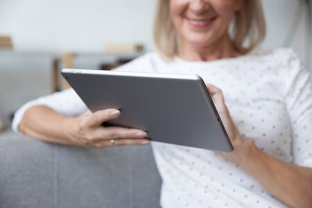 Smiling mature woman using computer tablet apps, holding electronic device in hands close up, positive old female surfing internet, chatting or shopping online, reading news in social networkの写真素材