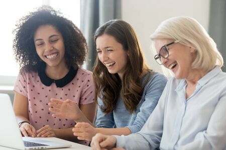 Diverse office workers sitting at desk watching funny video take break from work, mature young caucasian biracial colleagues laughing received humorous prank in internet, have fun at workplace conceptの写真素材