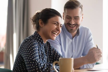 Happy young indian business woman laughing at funny joke with friendly colleague in office, cheerful diverse multi ethnic coworkers team having fun talking working together at break sit at deskの写真素材