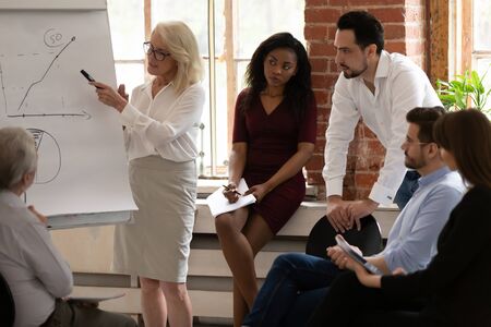 Middle-aged confident businesswoman stand talk making flip chart presentation for multiracial...
