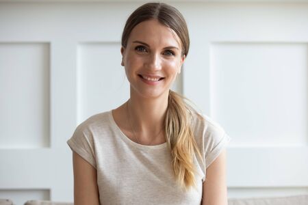 Head shot portrait of 30s woman sitting on sofa smiling looking at camera makes video call, distance...