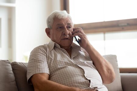 Elderly man in casual clothes seated on couch in living room holding mobile phone talking having...