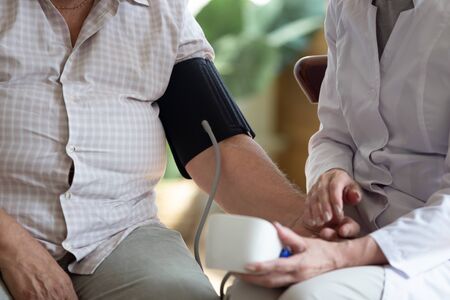 Seated indoors on couch caring nurse measures 70s patient blood pressure using sphygmomanometer...