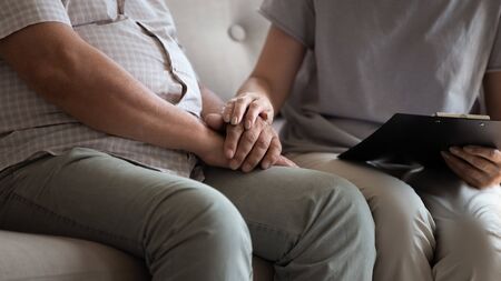 Close up conceptual image nurse holding clipboard and touch hands of patient talking sitting together on couch with elderly man, professional helpful caregiver comforting senior male at nursing homeの写真素材