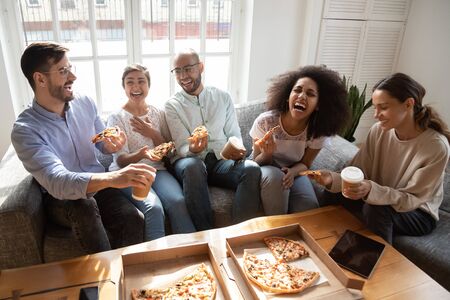 Happy diverse friends eating pizza, having fun together at home party, sitting on couch, laughing at joke, pleasant conversation, enjoying Italian junk food at meeting, satisfied customersの写真素材