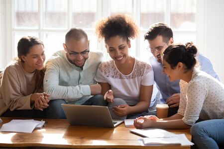 Happy diverse friends using laptop together, looking at screen, having fun, sitting on couch in...