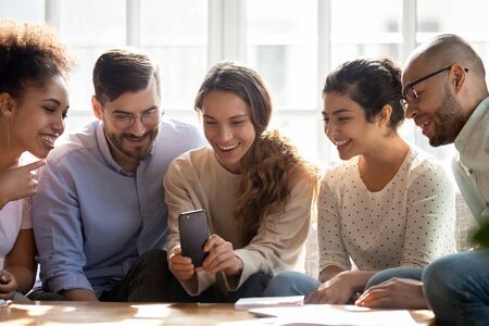 Smiling diverse friends using phone, sitting on couch, looking at screen, watching funny video in...