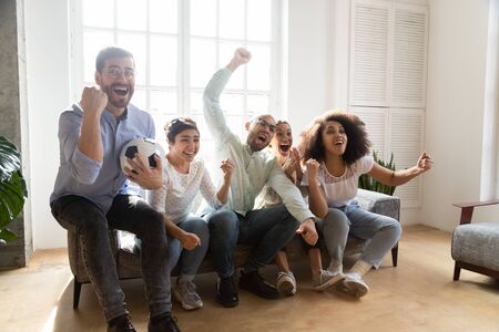 Excited diverse friends watching match on tv together, football fans celebrating victory goal, sitting on couch at home, screaming, supporting favorite soccer team, win sport bet, successful gameの写真素材