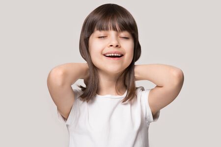 Calm happy little preschooler girl in white t-shirt isolated on grey studio background stretching...