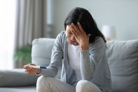 Frustrated unhappy mixed race woman sitting in living room, holding quick plastic test, worrying...
