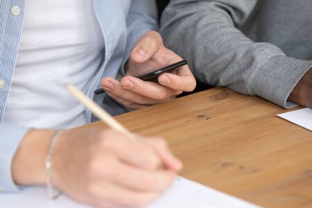 Close up diverse classmates sit at classroom desk, european guy writing info read on smartphone...