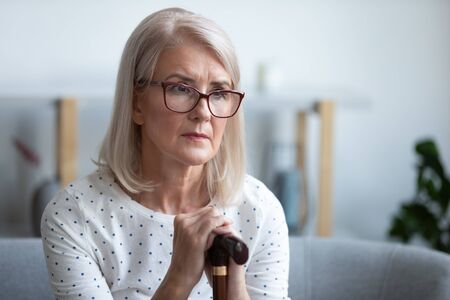 Sad lonely thoughtful pensive old mature woman hold cane stick sit alone on sofa, upset poor...