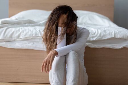 Front view upset young mixed race girl leaning back to bed sitting on floor, suffering from relations break up, betrayal, divorce. Depressed biracial woman feeling desperate, needs psychological help.の写真素材
