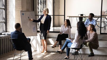 Serious middle aged businesswoman mentor coach corporate leader pointing on flip chart give...